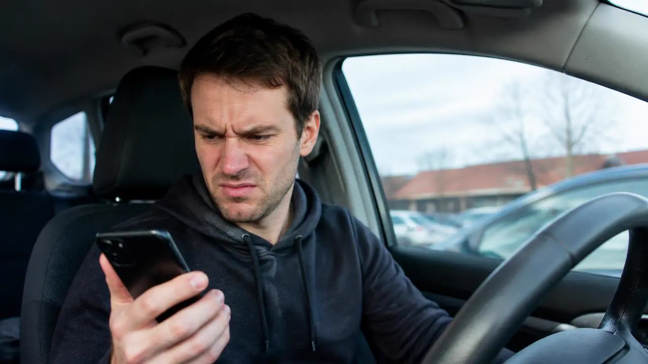Mann im geparkten Auto schaut nach dem Mailbox-Signalton genervt auf sein Smartphone